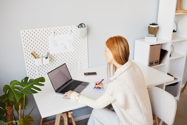 Ritratto ad alto angolo di giovane donna dai capelli rossi che usa il computer portatile o studia al minimo poliziotto sul posto di lavoro a casa...