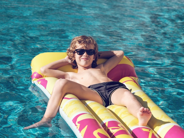 High angle of positive blond kid in shorts and sunglasses holding hands behind head and smiling while relaxing on inflatable float in swimming pool on resort in Mallorca, Spain