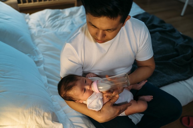 high angle shot loving Taiwanese father is holding and giving his newborn daughter bottled milk at bedside during bedtime in a dark bedroom at home.