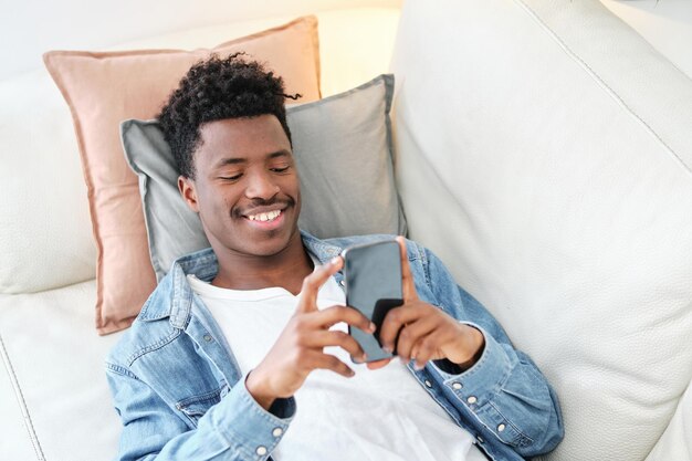 High angle smiling African American male in denim jacket browsing modern mobile phone and lying on comfortable sofa