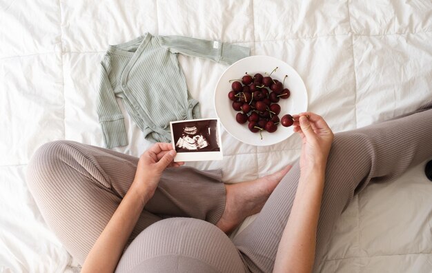 High angle top view of unrecognizable woman sitting on bed looking at ultrasound photo eating black