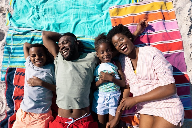 Высокий угол обзора афроамериканских родителей и детей, смеющихся, лежа на полотенцах на пляже. Без изменений, семья, вместе, детство, пикник, отдых, отдых, наслаждение и летняя концепция.