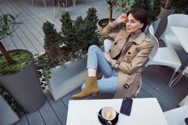 Photo high angle view of attractive elegant woman in blue denim jeans and beige eco leather coat relaxing at coffee break in outdoor cafeteria and looking dreamily to the side