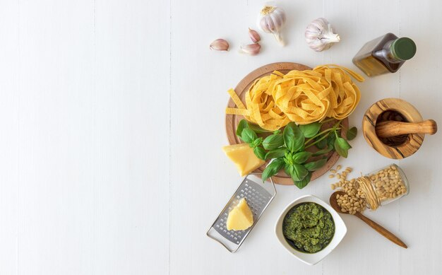 High angle view of basil pesto with ingredients and raw pasta on white wooden table background