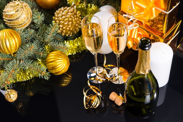 High angle view of champagne on shiny black surface with copy space and decorated evergreen branches, white candles and gold wrapped gifts