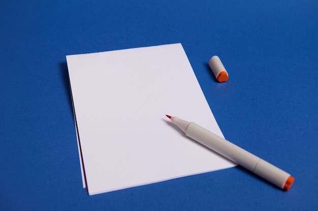 High angle view of a composition with watercolor marker or felt-tip pens on white blank sheet of paper with copy space for text isolated on blue background.