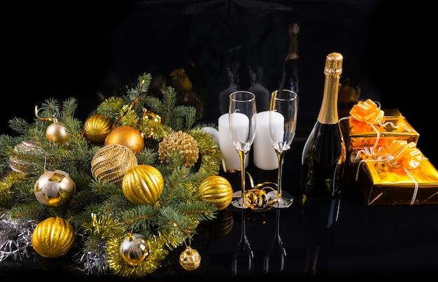 Праздничный натюрморт под высоким углом - бутылка шампанского с элегантными бокалами на черном фоне с подарками, свечами и вечнозелеными деревьями, украшенными елочными шарами и мишурой
