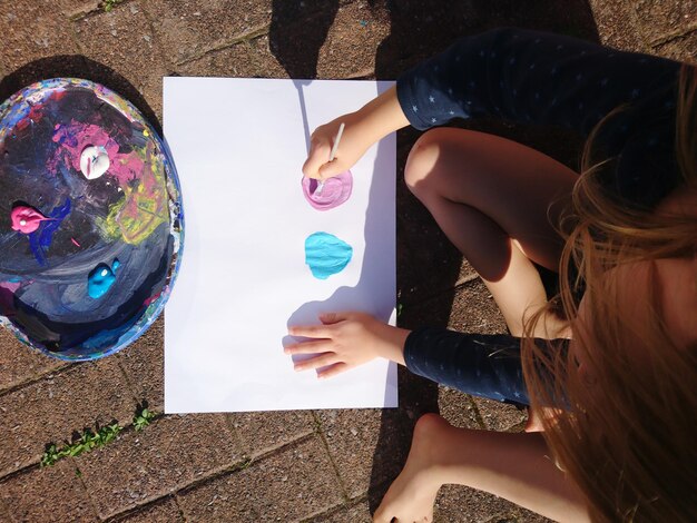 Photo high angle view of girl painting on paper while sitting at footpath