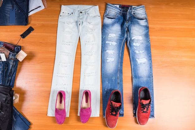 High angle view of his and hers distressed and torn blue jeans\
laid out flat on wooden surface with coordinating shoes - stylish\
pink loafers and red sneakers