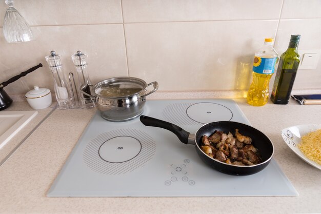 High Angle View of Kitchen Stove Top with Meat Frying in Pan and Pasta Boiling in Pot - Counter is Scattered with Other Ingredients for Meal Preparation