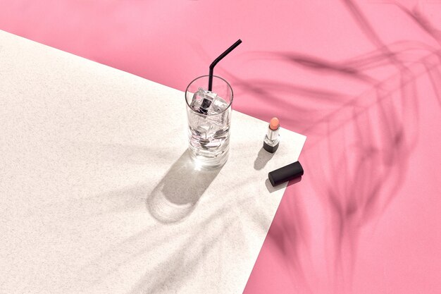 High angle view of lipstick and drinking glass with ice on table pink and white background with shad...