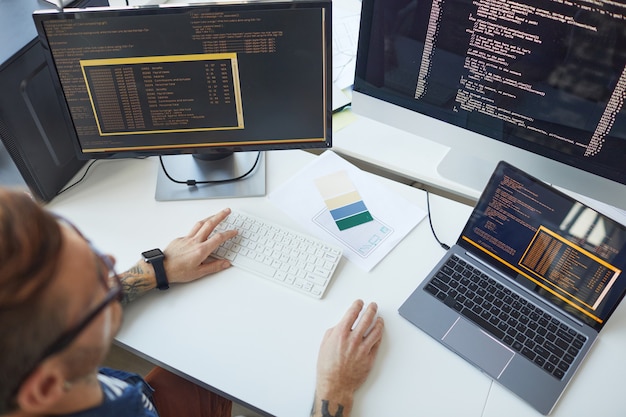IT開発者のオフィスで複数のコンピューターを使用しながらコードを書く男性プログラマーのハイアングルビュー、コピースペース