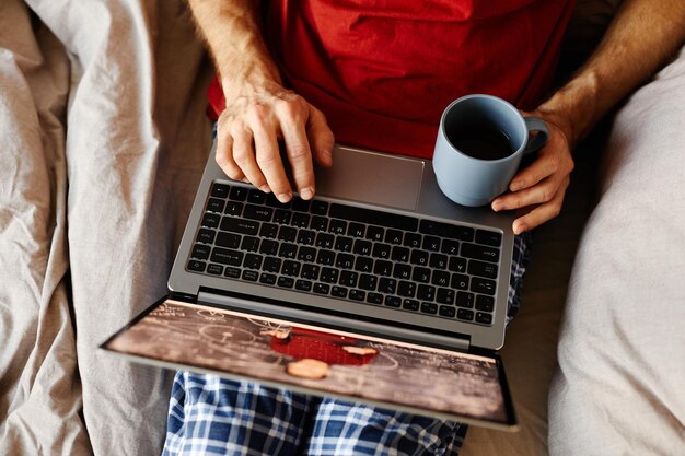 Photo high angle view of man watching video on laptop and drinking coffee while resting on his bed