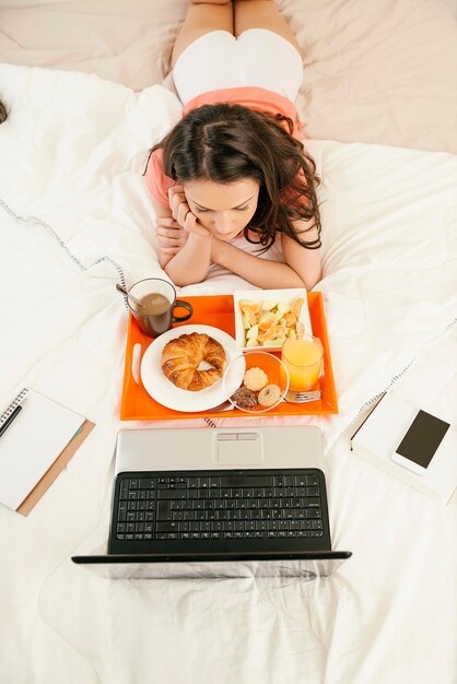 写真 ラップトップを見ながらベッドに横たわって朝食を食べている女性のハイアングルビュー