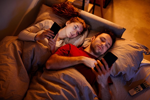 Высокий угол обзора молодой пары, использующей гаджеты перед сном, лежащей на кровати в спальне