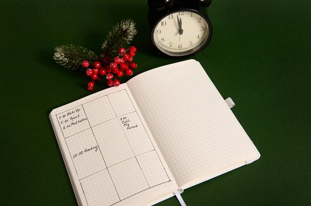 High angle view of an opened page of notepad with plans for day, alarm clock and snowy branch of holly Christmas plant isolated on green background