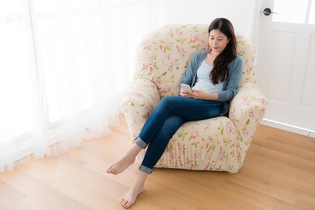 모바일 스마트폰으로 온라인 정보를 검색하고 창가 소파 의자 앞에 앉아 있는 웃고 있는 젊은 여성의 높은 각도 보기 사진.