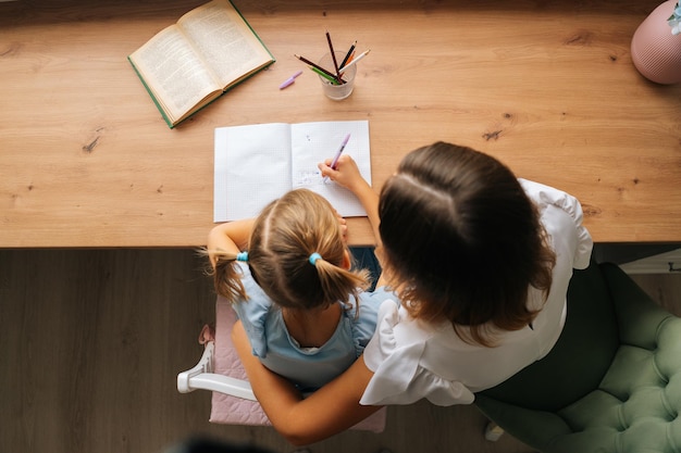 High-angle view of primary daughter doing homework writing in copybook with young mother sitting at home table. Top view of female tutor teaching elementary girl helping with lesson.