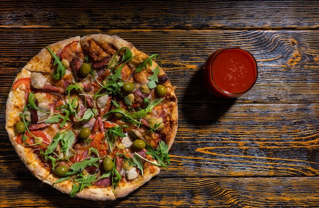 High angle view of tasty pizza with variety of toppings and cheese on wooden table near a glass with tomato juice