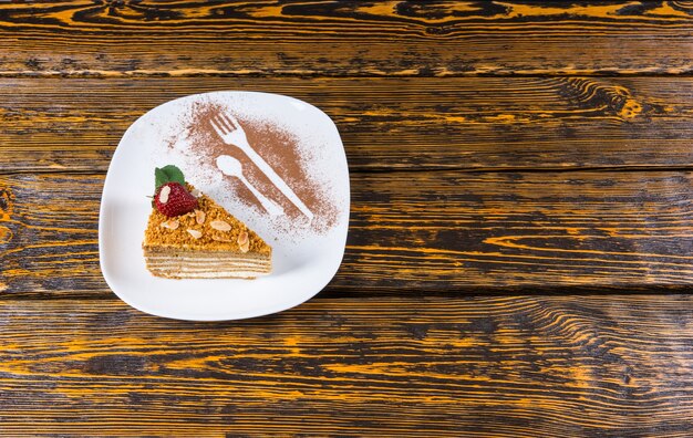 Elevato angolo di visione del cuneo triangolare di torta a strati guarnita con fragole e menta servita su un piatto bianco con utensili spolverati di cacao sulla superficie del tavolo in legno con spazio di copia