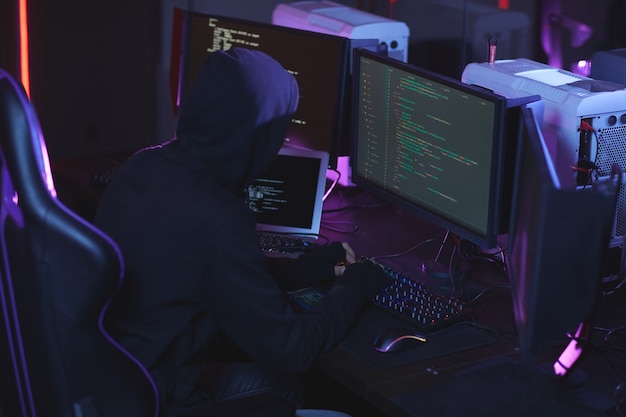 High angle view at unrecognizable cyber security hacker wearing hood while working on programming code in dark room, copy space