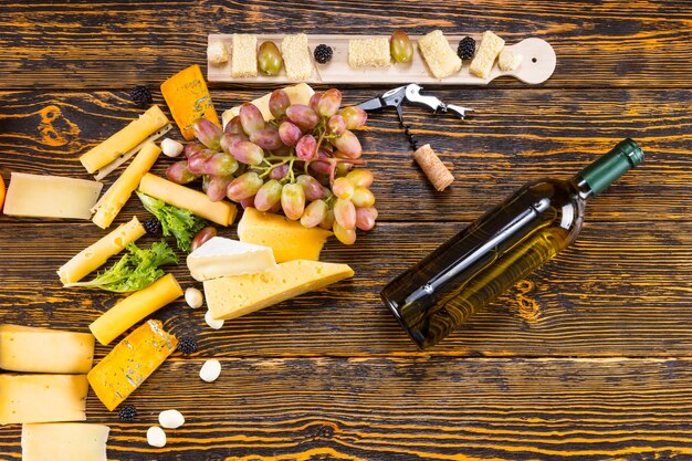 High Angle View of Variety of Gourmet Cheeses Scattered on Rustic Wooden Table with Grapes and Bottle of White Wine