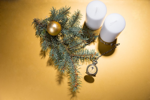 High angle view of white pillar candles on golden yellow background with evergreen branch, gold ball and antique pocket watch with copy space and spot lighting