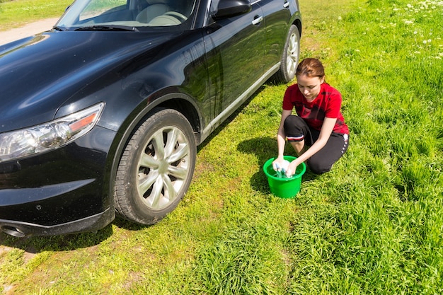 青い空と明るい晴れた日に石鹸スポンジを絞り、緑のフィールドで黒の高級車を洗う緑のバケツを持つ女性のハイアングルビュー