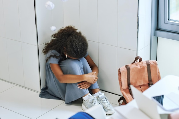 High angle view of young black girl bullied in school and sitting on floor with headphones