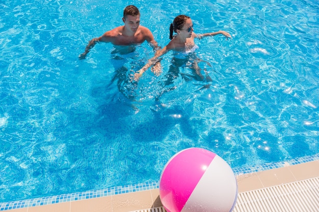 Elevato angolo di visione di una giovane coppia in piedi nel profondo del torace nella piscina del resort con pallone da spiaggia a strisce sul ponte della piscina