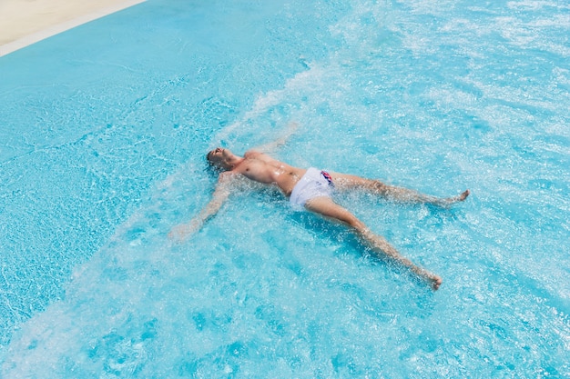 夏の晴れた日にプールの浅い波で腕と脚を広げて背中に横たわって水泳パンツを着ている若い男のハイアングルビュー
