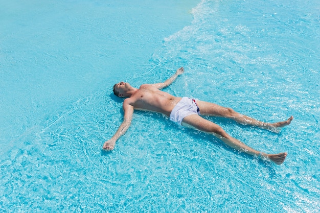 High Angle View of Young Man Wearing Swim Trunks Lying on Back with Arms and Legs Splayed in Shallow Waves of Swimming Pool on Sunny Day in Summer