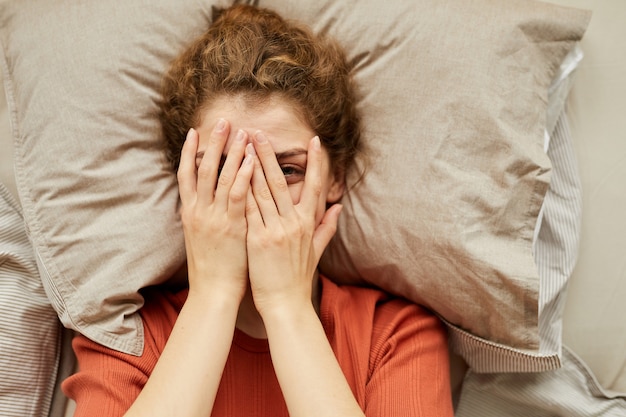 Photo high angle view of young woman looking at front while covering her face with hands while resting in bed