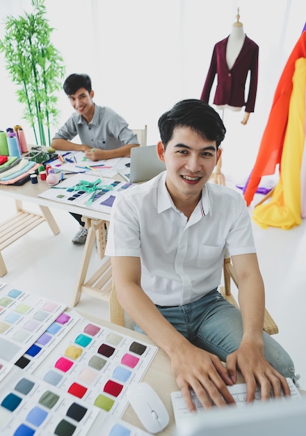 High angle of young Asian man smiling at camera and typing on computer keyboard while sitting at table with fabric samples near coworker in atelier