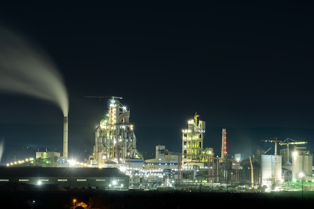 Высокая бетонная конструкция цементного завода с башенным краном и дымовой трубой в ночное время. Концепция промышленного производства и загрязнения воздуха.