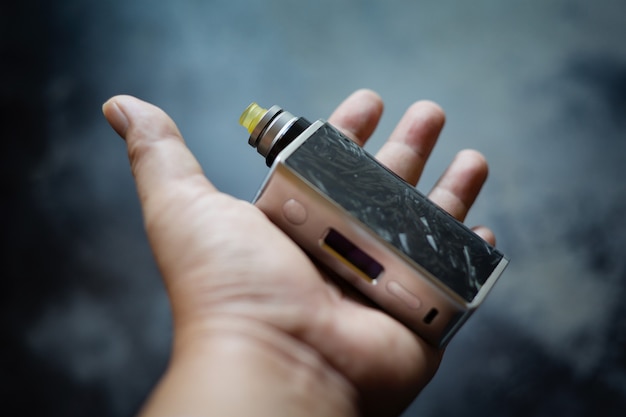 High end black carbon fiber in clear resin regulated box mods with rebuildable dripping atomizer and ultem drip tip in hand on dark grey texture background, vaporizer equipment, selective focus