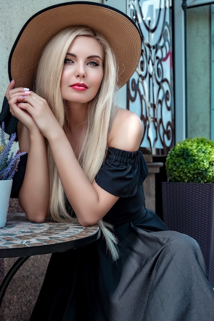 High fashion portrait of a graceful woman in an elegant straw hat and dress that sits in a cafe at a table