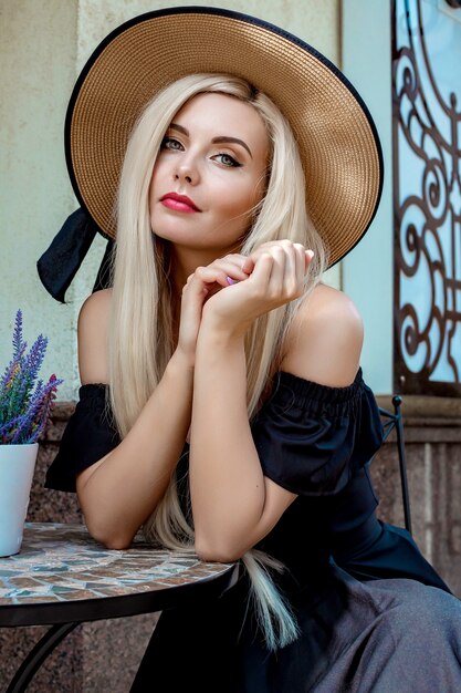 High fashion portret van een sierlijke vrouw in een elegante strooien hoed en jurk die in een café aan een tafel zit