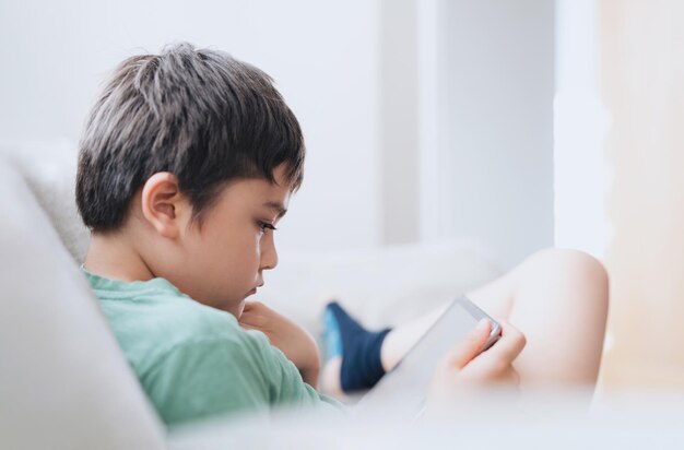 Photo high key portrait young boy playing game on tablet sitting on sofa with light shining from window kid playing games on internet cinematic portrait child doing homework online at home