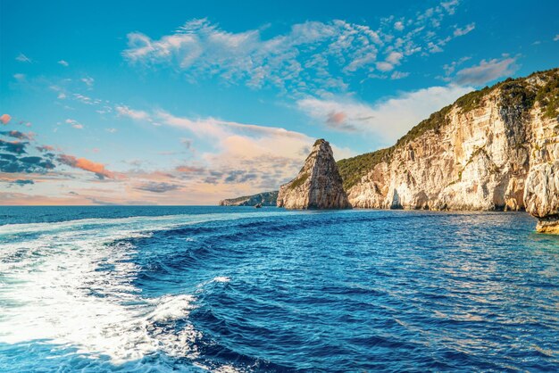 High layered lightcolored stoned rocky cliffs covered with plants on Corfu island sea beach with clear blue water under cloudy sky at sunset