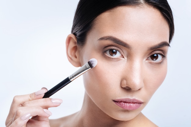 High-quality brush. The close up of a beautiful dark-haired dark-eyed woman holding a makeup brush near her face while posing