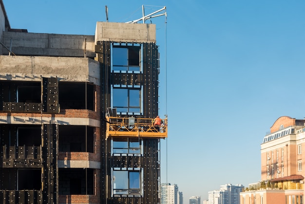 高層建設工事、ファサードを操作するクレードルの建設現場作業員、高層ビルの外で作業するビルダー用の吊り下げられたクレードル、建物の建設。