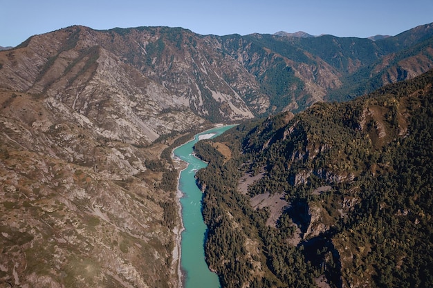 A high rocky mountains under a blue sunny sky and a narrow clear turquoise pond from a bird's-eye view.