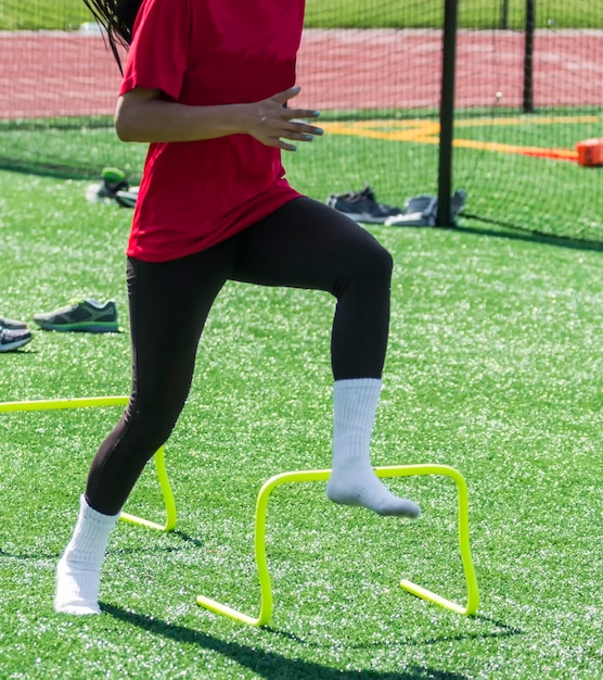 A high school girl is performing speed drills with no shoes on over yellow mini banana hurdles