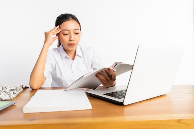 High school girl sitting at desk with laptop and holding book with thinking expression on isolated b