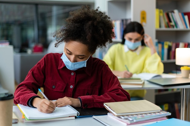 High school student wearing protective mask taking notes from book for her study Young woman sitting at desk at the background Pandemic concept