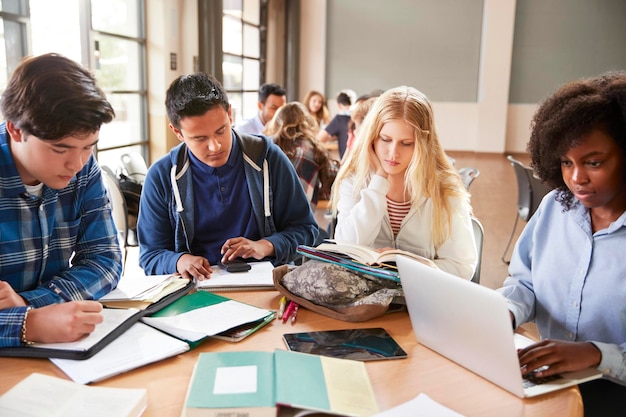 High School Students Using Laptops And Digital Tablets Working With Female Teacher At Desk