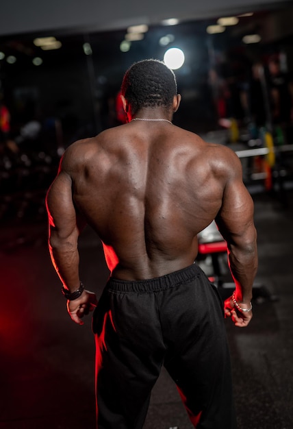 High and shirtless man poses with very muscular back to the camera. African american bodybuilder with perfect body. Dark gym background.