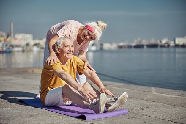 Un pensionato di buon umore che esegue un piegamento in avanti seduto sul tappeto assistito dalla sua allegra moglie