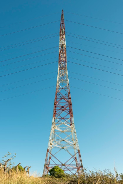 Стальная башня высокого напряжения, окрашенная в белый и красный цвета, с электрическими кабелями и сухой растительностью у основания.